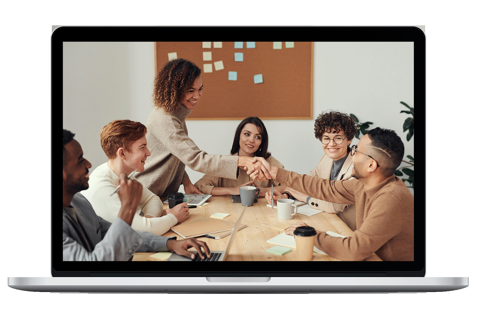 Image of a laptop computer with its screen displaying a group of six people in a meeting room. Two individuals are shaking hands across a table, while others look on and smile. The table has notebooks, coffee cups, a laptop, pens, and sticky notes, with a corkboard and plant in the background.
